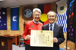 赤い服を着たジェニー市長と背広姿の高岡正人総領事が一緒に感謝状を持って記念撮影をしている写真
