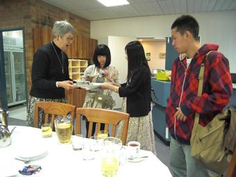 飲み物などが置かれたテーブルの横で日本人の女子学生が、外国人の女性に皿を手渡している写真