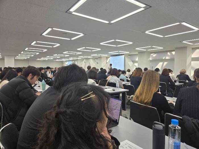 大きなホールで大人数の受講者がテーブルで座り、講義を受けている様子