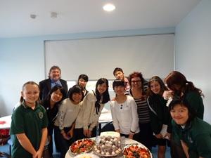 白い丸テーブルにたくさんの食べ物が並べられその周りを12人の外国人と日本人が並んでいる写真