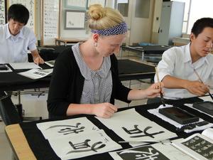 髪をアップにまとめたリズモー市の女子学生が、日本人の男子学生の隣に座り書道で漢字を書いている写真