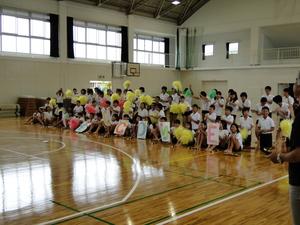 体育館で黄色や緑のボンボンを持った小学生たちが横3列に並んでいる写真