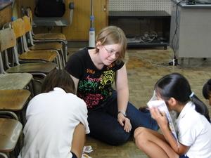 リズモー市の女子学生が床に座り、教科書で顔を少し隠している小学生の女の子に話しかけている写真