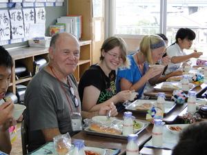 リズモー市の女子学生2人と関係者の男性が、机を並べて小学生達と一緒に給食を食べている写真