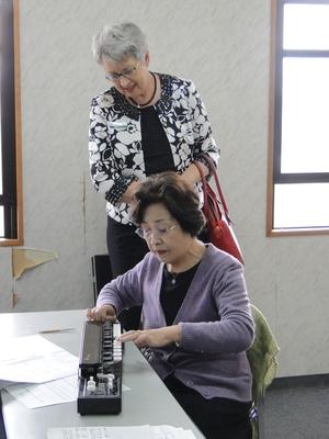 テーブルに置かれた大正琴を演奏している日本人女性を後ろから見学しているジェニー・ドウェル市長の写真