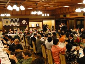 リズモー市の旗と大和高田市の旗が掲げられた広い室内で大勢の参加者達が席に座り手に枡を持って乾杯をしようとしている歓迎夕食会の様子の写真