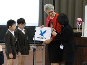 男子生徒2人が青い鳥の絵が描かれた紙を持ったジェニー・ドウェル市長の横に立ち、黒のスーツを着た女性がジェニー・ドウェル市長にマイクを向けている写真