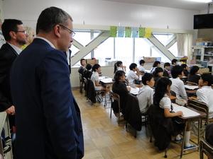 学校の子供たちの授業を部屋の後方から見学している外国人男性2人の写真