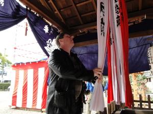 赤と白い布が神社の天井から垂れ下がっている白い布をキャメロンさんが両手で握っている写真