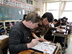 教室で生徒たちと机を並べ、女性に教わりながら書道をしているキャメロンさんの写真