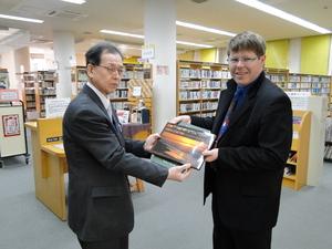 図書館内で、眼鏡をかけた日本人男性に2冊の本を手渡しているキャメロンさんの写真