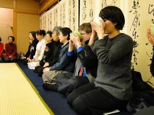 和室の壁際に洋服や、着物を着た女性達と並んで座り、お茶を飲んでいるキャメロンさんの写真