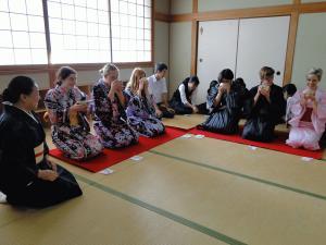 体育館で先生の指導を座って聞いているリズモー市からの生徒とバトミントンのコートで並んで座っている他の生徒達の写真