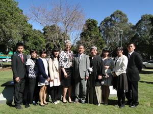 晴れた日の屋外で年配の外国人女性を中心に正装した学生と年配の日本人男性が並んで記念撮影をしている写真