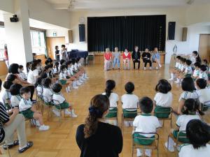 幼稚園児が椅子に座りリズモー市からの学生6名を紹介している先生の話を聞いている写真