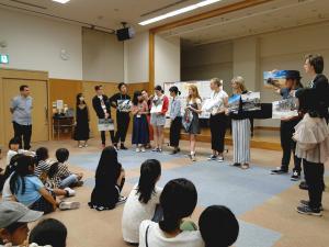 正面に外国人の日人達が並び1人の女子がマイクで説明をしているのを座って聞いている参加者の子供たちの写真