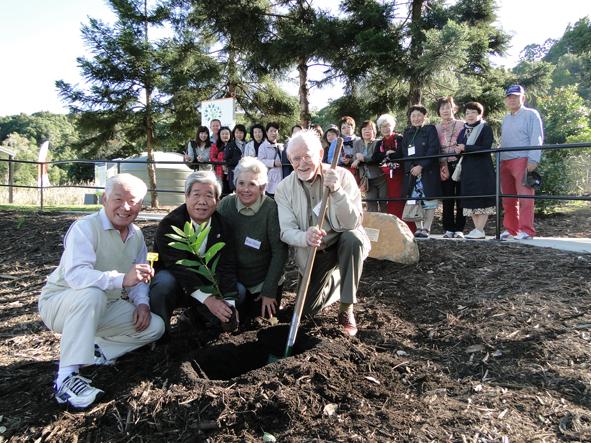 フェンスの奥で沢山の方が見守る中市長が木の苗を持ち、オーストラリア人女性、スコップを持った男性と記念植樹をしている写真