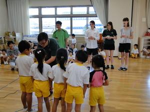 たくさんの園児が座っている中、5名の園児にリズモー市の1人の男子学生が腰を低くして会話をしている様子を4名の学生が後ろで見ている写真