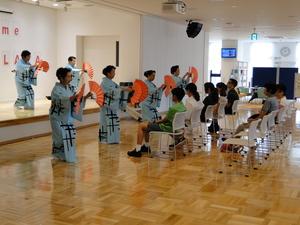 椅子に座るリズモー市の学生達のすぐ目の前でオレンジの扇を2枚使い踊りを披露している7名の方々の写真