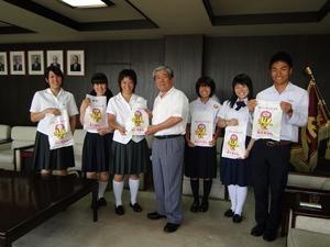 1人の女子学生が市長からマスコットキャラクターのみくちゃんが絵が描かれた袋を手渡され、その両側にみくちゃんが絵が描かれた袋を持って一緒に記念撮影をしている派遣学生達の写真