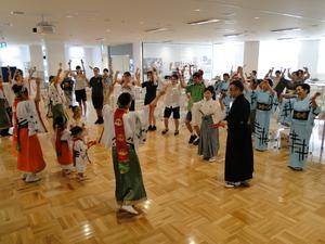 前列で鳴子を持ち両手をあげ踊りを教えている人達の真似をしているリズモー市の学生達と他の参加者達全体を写した写真