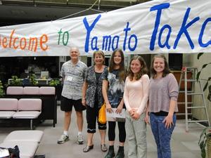 オレンジや、緑、青文字で「Welcome to Yamato Takada」と書かれた横断幕の前で記念撮影をしているリズモー市の女子学生3人、関係者の女性と男性の写真