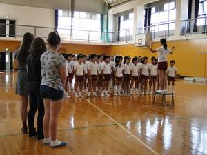 体育館に小学生が並んで歌を歌っている様子を見ているリズモー市の女子学生3人の写真