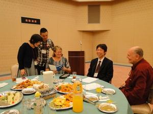 料理がのった丸テーブルに座るコリーンさんとリックさん夫妻と会話している3名の方の写真