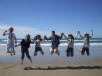 波打ち際で1人の外国人の男性と5人の日本人学生全員で手をつないでジャンプをしている写真