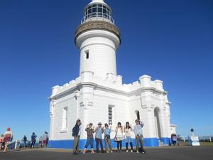 白い大きな灯台の前に並んで記念撮影をしている7人の学生の写真