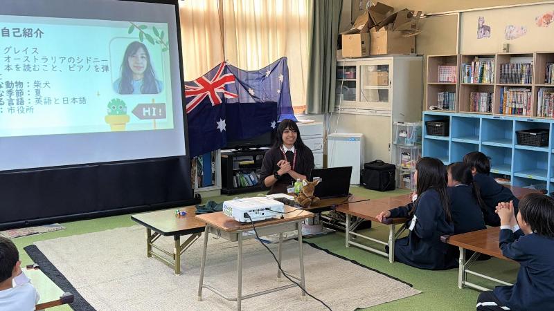 女性が机の前に座り、出前講座を行っている。子どもたちは前を向いてプロジェクターの画面を見ている。背景にはオーストラリアの旗が貼られている。