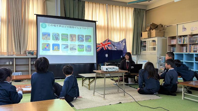 女性が机の前に座り、出前講座を行っている様子。子どもたちが前に向き、画面を見ている様子。背景にオーストラリアの旗が貼ってある。