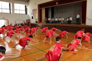 赤いお揃いの法被姿の生徒たちの踊りを体育館の舞台上から見学しているリズモー市の学生たちの写真