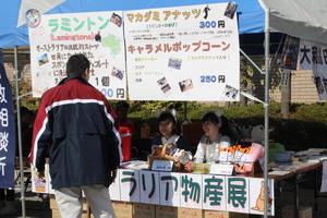 ラミントン、マカダミアナッツとキャラメルポップコーンの値段と紹介が書かれた用紙をテントに貼り接客をしている女子学生の写真