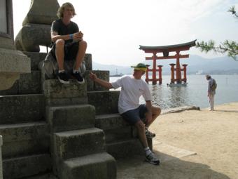厳島神社の海の中にある鳥居が後方に見える石段に腰かけている2人の外国人男性の写真