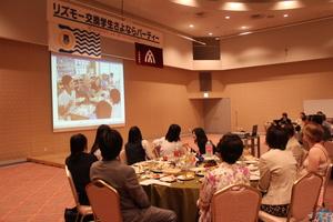 「リズモー市学生さよならパーティー」と書かれた横断幕が掲げれらた広いホールの前方に映し出された映像を見ている参加者達の写真