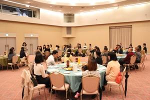 参加者の人達が丸テーブルに座っている部屋全体を写した写真