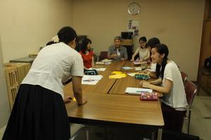 机をくっつけて座っている人達の中で一人の女子学生が立っている研修の様子を写した写真