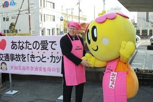 屋外で、ピンクの三角巾とピンクのエプロンを付けたキャメロンさんとピンクのエプロンを付けたマスコットキャラクターのみくちゃんが握手をしている写真