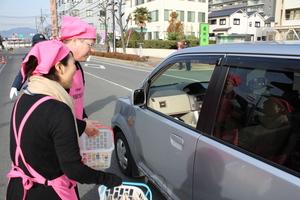 ピンクの三角巾、ピンクのエプロンを付けた女性とキャメロンさんがそれぞれ手に小さなかごを持ち、助手席側の窓が開いた車に話しかけている様子の写真