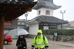 黄色の蛍光色のジャケットを着てジャケットのフードを被ったキャメロンさんと傘をさした女性が、雪が降る中を歩いている写真