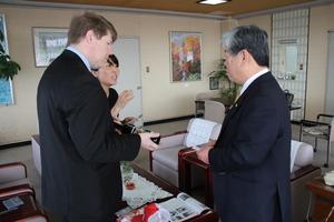 キャメロンさんが手にワインを持ち、吉田市長に渡そうとしている写真