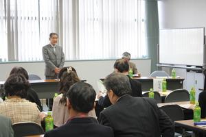 右側にホワイトボードが置かれた室内で、前方で立っている男性の話を聞いている参加者達の様子を後ろから写した写真