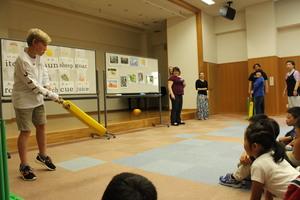 外国の男子学生が黄色のバットのようなものを持ちボールを打とうとしている所を子供達が見ている写真