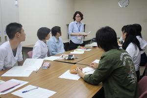 向き合わせて並べられた長机の奥側に3名、手前に3名の派遣学生が座り、長机の奥で首からネームタグを下げ立っている女性が学生達に話しかけている様子の写真