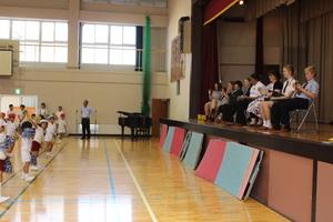 小学生が踊りを披露している様子を体育館舞台上で椅子に座り拍手をして見ている派遣学生達の写真