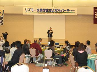 「リズモー交換学生さよならパーティー」と書かれた幕が掲げられた広いホールで、席に着いた参加者達が前方に立っている男性の方を見ている写真