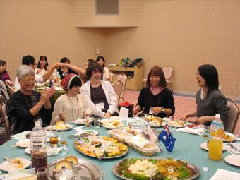 料理が並べられた丸いテーブルに日本人女性2人と外国人3名が座り談笑している写真