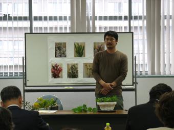 植物の写真が貼られたホワイトボードの前に立っているフローリストBee 長岡純一さんの写真