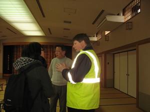 バックパックを背負った日本人女性、蛍光色の反射テープが付いたジャケットを着たキャメロンさん、日本人男性が広い和室内で立って話をしている写真